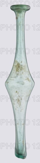 Unguentarium with an elongated belly and a long neck, 3rd–4th century AD, unknown type of site/place of origin, vessel, height 36 cm, width 7 cm, made in an unknown eastern workshop., A tall, narrow glass object with a long neck and a flared lip, widening into a symmetrical, diamond-shaped midsection and tapering again toward the base; the glass is pale greenish-blue and translucent with several cloudy patches, surface scratches, and areas of brownish discoloration or residue scattered across the midsection and neck; the lip is slightly darker green at the rim, and the base end shows a small irregularity and darker green tint.