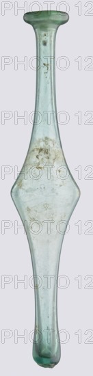 Unguentarium, unknown eastern workshop, Goluchów Greater Poland Voivodeship — with an elongated belly and a long neck, 3rd–4th century AD, vessel, height 36 cm, width 7 cm, A slender elongated glass object with a narrow neck that widens into a flattened, diamond-shaped midsection and tapers again toward a rounded end, shown upright against a pale background; the glass is translucent with a greenish-blue tint and shows several cloudy areas, surface scratches, and patches of residue in a brownish-beige color scattered on its surface, with slight variations in thickness creating subtle color intensity differences and a few small bubbles or irregularities visible within the glass.