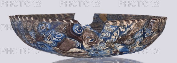 Bowl, Egyptian workshop; mosaic glass, glass vessel with floral patterns. Bowl with a flat bottom whose walls widen evenly toward the top then narrow at the rim; cut flat at the top. Made of thick glass using the mosaic technique; floral patterns in blue, purple, yellow, green, and white. Type: Isings form 1. Semicircular bowl; sagging on the form; 1st century, Roman Empire 27 BC–476. Vessel; height 3 cm, width 13.5 cm., A shallow, broken bowl with an irregular rim showing a jagged gap, decorated with swirling, concentric patterns in shades of blue, brown, beige, and white; the surface shows areas of glossy and matte texture with worn and rough patches, and the interior and exterior share the same patterned design, all set against a pale gray background with a subtle reflected shadow beneath.
