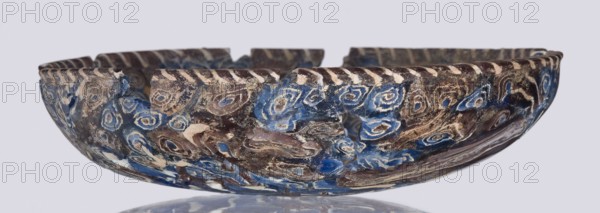 Bowl, Egyptian workshop; mosaic glass; floral patterns in blue, purple, yellow, green, and white. Bowl with a flat bottom whose walls widen evenly towards the top then narrow at the rim, cut flat at the top. Made of thick glass using the mosaic technique. Type: Isings form 1. Semicircular bowl; sagging on the form; 1st century, Roman Empire; vessel; height 3 cm, width 13.5 cm., A shallow bowl with a scalloped rim sits against a pale gray background, showing a marbled surface of swirling blue, brown, cream, and white tones. The exterior displays a pattern of rounded, eye-like swirls and irregular patches where the colors blend, with areas of rough texture and small flakes. The rim has a repeating diagonal stripe effect in cream and dark brown, and there are a few notches cut into the rim. The inside edge echoes the exterior colors and marbling, and the bowl rests on a reflective surface that mirrors the blue and brown hues.