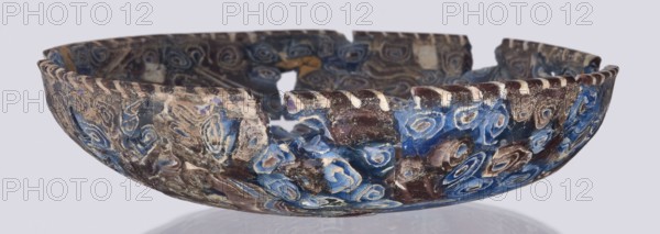 Bowl, Egyptian workshop; mosaic glass; glass vessel decorated with floral patterns in blue, purple, yellow, green, and white. Bowl with a flat bottom whose walls widen evenly toward the top then narrow at the rim; cut flat at the top. Made of thick glass using the mosaic technique Isings form 1. Semicircular bowl; sagging on the form; 1st century, Roman Empire. Vessel; height 3 cm, width 13.5 cm., A broken shallow bowl with a scalloped rim showing a pattern of swirled, concentric designs in shades of blue, brown, beige, and white, with areas of rough, weathered surface and chips exposing inner textures; the bowl rests on a reflective surface and the background is a pale gray.