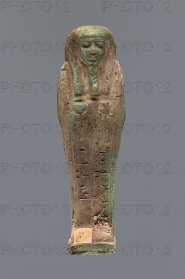 Ushabti, Hor-em-saf ca. 664–525 BC. Mummy-shaped figurine made of green faience. Stocky build, standing on a base, supported at the back by a pillar/pilaster. Arms crossed on the chest; from under the shroud only the hands, clenched into fists, are visible. Right hand holds a hoe, left holds a pickaxe. A basket on a single string is slung over the left shoulder. Engraved tools. Smooth, three-part wig; straight, braided beard. Large eyes with downcast eyelids, prominent mouth with a slight smile, wide nose. Engraved inscription in two columns on the front of the legs: Shd Osiris Hor-em-saf born of T-ate-iset. 664–525 BC; Late Period, 26th Dynasty, Egypt. Type: shabti. Height 10.6 cm; width 3.2 cm., A small standing human-shaped figurine with a rounded head and stylized facial features, depicted with a long headdress that frames the face and falls along the sides of the body; the surface shows a weathered, rough texture with areas of greenish patina and brownish-beige tones, and the fron...