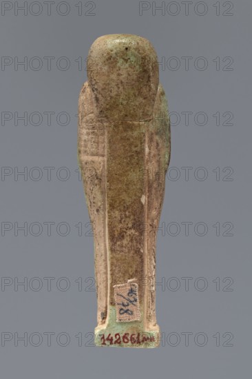 Ushabti, Hor-em-saf ca. 664–525 BC. Mummy-shaped figurine made of green faience. Stocky build, standing on a base, supported at the back by a pillar/pilaster. Arms crossed on the chest; from under the shroud only the hands clenched into fists are visible. Holds a hoe in the right hand and a pickaxe in the left; a basket on a single string hangs over the left shoulder. Engraved tools. Smooth three-part wig, straight braided beard. Large eyes with downcast eyelids, prominent mouth with a slight smile, wide nose. Engraved inscription arranged in two columns on the front of the legs reading: Shd Osiris Hor-em-saf born of T-ate-iset. 664–525 BC; Late Period, 26th Dynasty, Egypt. Type: shabti. Dimensions: height 10.6 cm, width 3.2 cm., A single elongated statuette viewed from the back, with a rounded top and a tapered rectangular body that flares slightly at the base; the surface shows worn and uneven texture with patches of light green, tan, brown, and darker brown mottling, and there is a ...