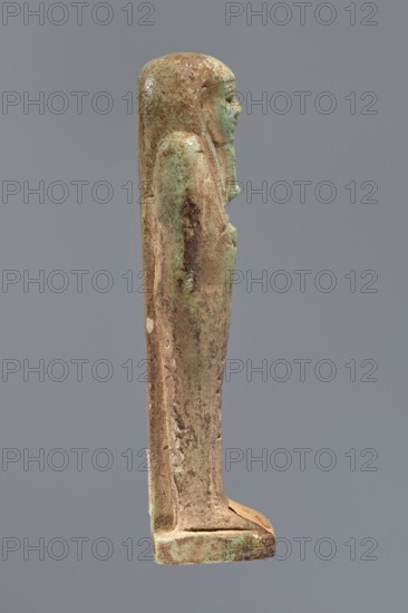 Ushabti, Hor-em-saf ca. 664–525 BC. Mummy-shaped figurine of green faience, 10.6 cm high, 3.2 cm wide. Stocky figure standing on a base, supported at the back by a pillar. Arms crossed on the chest; only the hands visible from under the shroud, clenched into fists holding a hoe right and a pickaxe left. A basket on a single strap is slung over the left shoulder. Engraved tools. Smooth three-part wig; straight, braided beard. Large eyes with downcast eyelids, prominent mouth with a slight smile, wide nose. Engraved inscription in two columns on the front of the legs: Shd Osiris Hor-em-saf born of T-ate-iset. 664–525 BC; Late Period, 26th Dynasty, Egypt., A slender upright statuette viewed in profile against a plain gray background, showing a standing figure with a smooth rounded head and a long straight body fused to a small rectangular base; the surface has irregular textures and areas of wear, with a mottled palette of pale green, light brown, and darker brown patches, and small spots...
