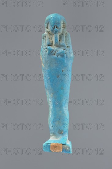 Ushabti, Mummy-shaped figurine, standing on a base, back supported by a pillar/pilaster. Arms crossed on the chest, right folded over the left. Carved hoes in both hands. On the back is a bag, slung over the left shoulder on a single string. On the head is a three-part, smooth wig, merging into a pilaster at the back. Long, straight beard, tapering downwards. Carelessly crafted facial features, bulging eyes, mouth in a slight smile. Mummy-shaped figurines, grave goods, wigs, hoes. 332–30 BC; Late Period–Ptolemaic period 27th–30th dynasty, Egypt. Shabti, height 9.5 cm., A small upright figurine in bright turquoise-blue with areas of lighter and darker blue and small spots of beige and brown, depicting a wrapped standing figure with a rounded head and simplified facial features, arms crossed on the chest, a few small pits and surface irregularities, and a rectangular base with a small worn label attached.