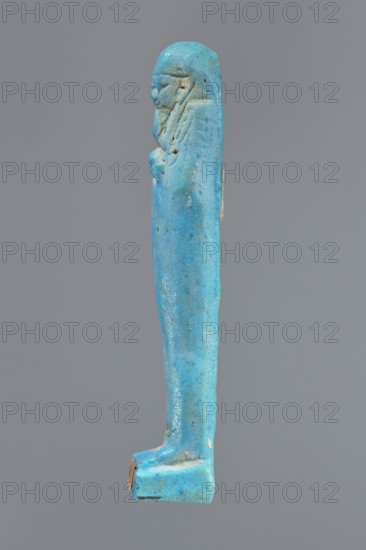 Ushabti, mummy-shaped figurine standing on a base, back supported by a pillar/pilaster. Arms crossed on the chest, right folded over the left. Carved hoes in both hands. On the back is a bag, slung over the left shoulder on a single string. On the head is a three-part, smooth wig merging into a pilaster at the back. Long, straight beard, tapering downwards. Carelessly crafted facial features, bulging eyes, mouth in a slight smile. 332–30 BC; Late Period–Ptolemaic period 27th–30th dynasty, Egypt. Shabti, height 9.5 cm., A slender vertical blue object resembling a standing figure with a rounded head and simplified facial features in profile, a suggestion of hair or a headdress running down the sides, a smooth elongated body tapering toward the feet, and a flat rectangular base; the surface shows variations in blue tones with some darker speckles and small areas where the blue is worn to reveal a brownish color, all set against a plain gray background.