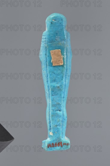 Ushabti, Mummy-shaped figurine standing on a base, back supported by a pillar/pilaster. Arms crossed on the chest, right folded over the left. Carved hoes in both hands. On the back is a bag slung over the left shoulder on a single string. On the head is a three-part, smooth wig merging into a pilaster at the back. Long, straight beard tapering downwards. Carelessly crafted facial features, bulging eyes, mouth in a slight smile. Materials: likely faience or similar; function: shabti grave figurine. Date: 332–30 BC, Late Period–Ptolemaic period 27th–30th dynasty, Egypt. Height: 9.5 cm., A small statuette in a bright turquoise-blue color shaped like a standing human figure with simplified arms and a rounded head, showing surface texture with some darker blue speckles and small irregularities, with a small rectangular patch of beige fabric attached to the front, all set against a plain gray background.