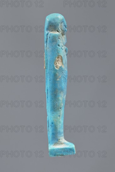 Ushabti, Mummy-shaped figurine made as grave goods, standing on a base with the back supported by a pillar/pilaster. Arms crossed on the chest, right folded over the left, holding carved hoes in both hands. A bag is on the back, slung over the left shoulder on a single string. Wears a three-part smooth wig merging into the pilaster at the back, and a long straight tapering beard. Carelessly crafted facial features with bulging eyes and a slight smile. Dated 332–30 BC, Late Period–Ptolemaic period 27th–30th Dynasty, Egypt. Shabti, height 9.5 cm., A slender vertical figurine in bright turquoise-blue with areas of lighter and darker blue and some small pale worn spots, shown in profile against a plain gray background; the figure has a rounded head and a suggestion of facial features, a long robe-like body with a few rough pitted areas and surface irregularities, and a flat rectangular base in the same blue tones.