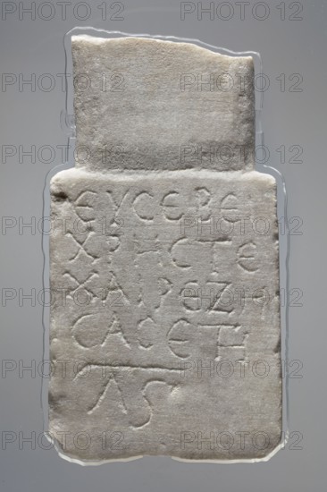 Epitaph of Eusebius, stele in the shape of two rectangular slabs of different widths placed narrower above the wider one. The lower slab has five lines of Greek text. 2nd–3rd century paleography. Sidon. Tombstone inscription in Greek. Height 24.5 cm, width 13.7 cm, depth 3.6 cm., A rectangular stone slab with an irregular top edge resting on a neutral background; the slab is a pale off-white color with subtle gray and beige variations and a slightly rough, weathered surface texture. The front face shows a group of shallow incised linear markings forming repeated shapes across the surface, and there are a few small chips and rounded wear along the slab edges. The background is a smooth gray with a faint darker gradient, and there is a thin clear outline around the slab where it meets the background.