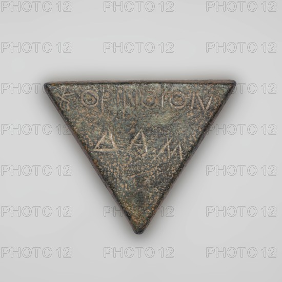 Weight 1/6 mina. Weight in the shape of an isosceles triangle. On the weight is a two-line inscription in Greek KORINTHION, below it DAM. 3rd century BC, Corinth. Weight; height 4.5 cm, width 5.5 cm, depth 0.7 cm, weight 73.4 g., A triangular metal object with a textured, weathered surface in shades of green, brown, and dark gray, showing areas of lighter tan speckling; the object has rounded edges and a slightly raised triangular profile, with a group of shallow incised marks visible on its surface; the background is a smooth, evenly lit pale gray.