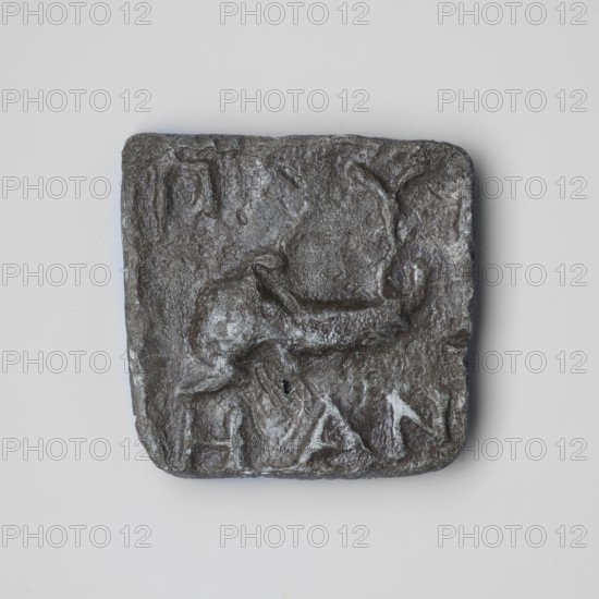 Weight 1/2 mina. Square weight with straight side edges, slightly protruding above the face. The face bears a plastic representation of a scorpion facing left, arranged horizontally in the central part. The relief contours are not distinct, blending with the background. Above the animal, in the corners, are the relief Greek letters GY, below EAN. Loss in the upper left corner. Mid-4th century BC, Byzantion, weight. Dimensions: height 5 cm, width 5 cm, depth 0.7 cm; weight 214.1 g., A square, textured object with rounded corners sits centered against a light gray background; the object is dark gray to charcoal with a rough, uneven surface showing raised and indented areas, including a few smooth shiny patches and some lighter gray highlights; the surface has irregular contours that suggest carved or molded forms and a few shallow depressions and ridges across its face.