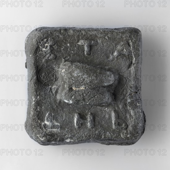 Square weight with diagonally cut side edges. The upper surface is decorated with a plastic representation of an astragalus. At the upper edge are horizontally arranged convex letters ETA, at the lower edge THb. A second stamp with an image of a sea turtle was impressed next to the astragalus. 6th century BC, Attica. Weight 2 trade mines, unknown Attic workshop. Weight: 1205 g; height 7.1 cm; length 6.9 cm; width 2.6 cm., A roughly square, dark grey object with an uneven, textured surface sits against a light grey background; the object has rounded corners and a raised, irregular central shape that projects from the flatish face, and there are a few shallow depressions and raised marks around the central form, with small patches of lighter grey scattered across the surface.