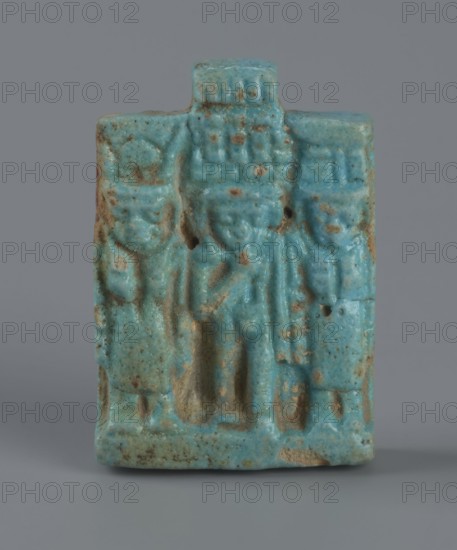 Amulet – plaque with a triad of deities: Harpocrates Horus, Isis, Nephthys. Amulet in the shape of a plaque with a triad of deities standing on a base: central figurine of Horus, Isis at left, Nephthys at right. Back of plaque flat; pendant above the upper edge. Date: 664–30 BC, Late Period–Ptolemaic Period probable. Country of find: Egypt. Material/function: amulet. Dimensions: height 3.3 cm, width 2.2 cm, depth 0.7 cm., A small rectangular turquoise-blue carved object with a slightly rough, speckled surface and patches of brownish discoloration, showing a raised central figure flanked by two smaller raised figures; the top has a short rectangular projection, and the lower edge is slightly worn with visible areas of exposed material; the background and base are a neutral gray.