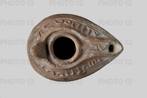 Oil lamp, deposit, Islamic art, Arabic, ceramics, Arabic ceramics, utility ceramics, Arabic inscriptions, Arabic language, everyday life in the Middle East. Oval lamp from a mold. Gently sloping arms with a Kufic inscription bismi l'lahi mina l'lahi = in the name of Allah, from Allah. A large oil opening surrounded by a convex shaft. A second outer shaft also surrounds the burner channel, on which another inscription one word is inscribed. Conical nozzle. Ring-shaped round base base-ring, in the center of which there are Arabic letters l, r, k?. 8th–9th century, Syro-Palestine. Oil lamp; height 4.4 cm, length 9.1 cm, width 6.4 cm., An oval, teardrop-shaped ceramic object viewed from above with a central circular opening and a narrow spout-like projection, featuring raised ridges and carved decorative grooves; the surface shows a weathered, rough texture with areas of erosion and patina. The visible colors include brown, reddish-brown, gray, and a few lighter beige tones.