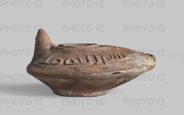 Oil lamp, Oval lamp from a mold. Gently sloping arms with a Kufic inscription bismi l'lahi mina l'lahi = in the name of Allah, from Allah. A large oil opening surrounded by a convex shaft. A second outer shaft also surrounds the burner channel, on which another inscription one word is inscribed. Conical nozzle. Ring-shaped, round base base-ring, in the center of which there are Arabic letters l, r, k?. Islamic art; Arabic ceramics; utility ceramics; Arabic inscriptions; Arabic language; everyday life in the Middle East. 8th–9th century, Syro-Palestine. Materials: ceramics. Function: oil lamp. Dimensions: height 4.4 cm, length 9.1 cm, width 6.4 cm., A weathered ceramic object rests on a neutral gray surface and background, showing a rounded base that flares outward into a wider midsection and tapers to a pointed protrusion at one end and a raised spout-like rim at the top. The surface texture is rough and pitted with visible chips and cracks, and there are shallow carved or impressed gr...