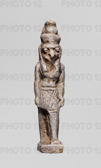 Amulet - god Horus; maker, Horus mythology; double crown iconography, double crown on the head; Egyptian deity; Late Period, 664–332 BC, Egypt; amulet; height 3.8 cm, width 0.8 cm, A small carved figurine of a standing humanoid with an animal-like face and a tall, layered headdress, shown against a plain light gray background; the figure is rendered in a weathered stone with a palette of gray, off-white, and brown tones, with darker brown areas accenting crevices and worn details; the figure stands on a rectangular base, wears a short skirt with vertical textured lines, and has arms held close to the sides; the surface shows pitting and uneven texture and the overall form is slender and elongated.