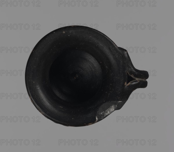 Black-finned oinochoe, unknown Attic workshop, oinochoe, Ancient Greece, ancient vessel, black-finned pottery, Black-finned oinochoe with a wide cylindrical foot directly transitioning into a wide, bulging body decorated with fluting. The body transitions into a wide neck without emphasizing the shoulders, ending in a wide, flat rim. The handle is made of two vertical rollers joined together, connecting the upper part of the body with the rim edge, to which it is attached with split rollers. 3rd century BC, Attica, vessel, A top-down view of a dark round vessel with a wide rim and a deep concave interior, predominantly black with small areas of worn brown and a patch of lighter scuffing, set against a flat neutral gray background; the vessel has a short dual-pronged spout on one side and visible surface texture and wear marks.