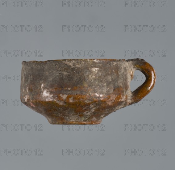 Miniature scoop with a flat bottom; walls widening diagonally to 1/3 then running vertically upwards; one vertical cylindrical vice., Origin: Greece. Type: vessel, scoop. Height 1.5 cm; width 2.5 cm., A small, worn ceramic cup with an integrated loop handle is shown against a plain grey background; the cup has an uneven, rounded body with a slightly narrower foot and a rim that appears irregular; its surface shows a mottled mix of brown and grey tones with patches of lighter, off-white areas and some darker spots, and the handle displays similar brown and grey coloration.