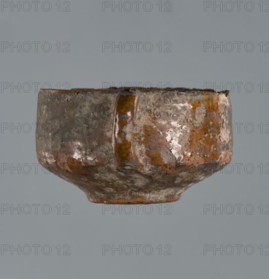 Miniature scoop with a flat bottom; walls widen diagonally to 1/3 then run vertically upward; one vertical cylindrical handle., origin Greece; vessel/scoop; height 1.5 cm, width 2.5 cm., A small rounded ceramic cup with a single loop handle, shown against a gray background, exhibiting a textured, uneven surface with worn glaze. The cup's visible colors include brown, rusty orange, gray, and patches of off-white, with glossy and matte areas where the glaze has chipped or weathered. The handle is attached near the rim and shares the same mottled coloration, and the cup's base tapers slightly inward.