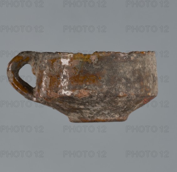 Miniature scoop with a flat bottom; walls widen diagonally to 1/3 then run vertically upwards; one vertical cylindrical handle/vice. Material unspecified. Origin: Greece. Type: vessel/scoop. Height 1.5 cm, width 2.5 cm., A small, worn cup with a handle is shown against a plain grey background; the cup surface appears rough and weathered with patches of brown, orange, grey, and hints of white, and the handle has similar brown and orange tones.
