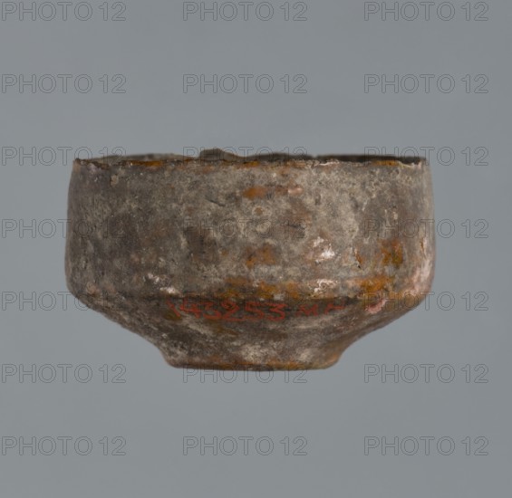 Miniature scoop with a flat bottom; walls widening diagonally to 1/3 then running vertically upwards; one vertical cylindrical vice., Place of origin: Greece. Type: vessel, scoop. Dimensions: height 1.5 cm, width 2.5 cm., A small, rounded bowl with a rough, weathered surface is shown against a plain gray background; the bowl's exterior displays a mottled mix of gray, brown, rusty orange, and patches of pale whitish discoloration, with an uneven rim and a slightly narrower base, and the overall texture appears pitted and corroded.