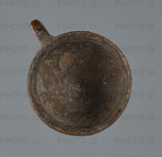 Miniature scoop with a flat bottom; walls widen diagonally to 1/3 then run vertically upwards; single vertical cylindrical handle., place of origin: Greece. Vessel/scoop; height 1.5 cm, width 2.5 cm., A top-down view of a round, shallow vessel with a small handle, showing a worn interior surface in shades of brown, dark brown, grey, and hints of orange, set against a uniform mid-grey background.