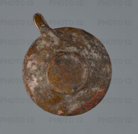 Miniature scoop with a flat bottom; walls widening diagonally to 1/3 then running vertically upwards; one vertical cylindrical handle., origin Greece; vessel/scoop; height 1.5 cm, width 2.5 cm., A round, flat object with a short handle projecting from one side, showing a rough, weathered surface with mottled areas in brown, rust, gray, and patches of green and pink, set against a plain gray background.