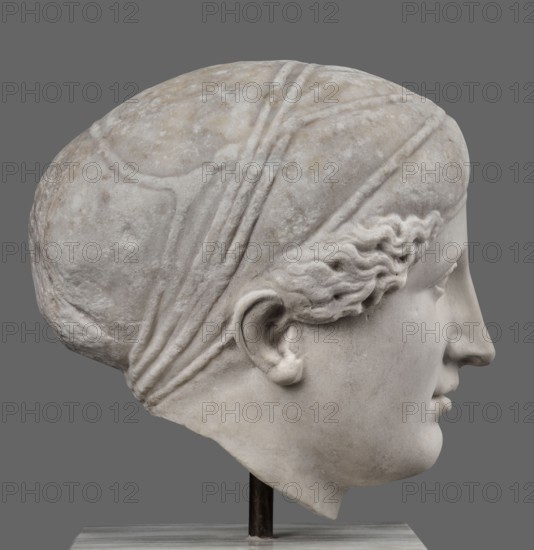 Head of Sappho Head of a Woman of the Kora Albani type, unknown Roman workshop. Sappho ca. 610–580 BC . Female portrait, Roman sculpture. Fragment of an ancient dowry or bust mounted on a modern bust. Numerous modern additions to details e.g., tip of the nose. The ancient part very good workmanship was originally part of a statue or bust herm?. Head slightly turned to the right, oval, idealised face. Hair covered with a headnet kekryphalos. Sculpture's original identified as contemporary to Phidias; dating possible by analogy to one of the Parthenon friezes. Type belongs to the Sappho/Kora Albani group, based on near-intact preservation. Front is very well-crafted; back is simplified and somewhat underdeveloped. Copy has several minor details altered compared to the original. Early 1st century, Roman Empire 27 BC–476. Sculpture, weight 30.5 kg., A marble sculpted head shown in profile against a gray background, with pale white and light gray stone tones and subtle beige discoloration; ...
