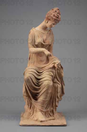 Figurine of a woman sitting on a rock with a flower in her hand. Woman sitting on a rock with a flower in her hand, towards which she bows her head. She is dressed in a thin chiton fastened at the shoulders and tied high under the breasts; a pleated cloak is thrown over her knees and left forearm. Hair is combed upwards. Flat quadrangular base. 4th century BC, Myrina. Figurine; height 31.5 cm, width 12.5 cm, depth 18 cm, weight 0.78 kg., A seated terracotta figure rendered in earthy tones of reddish-brown and beige sits on a rectangular base against a neutral gray background; the figure wears a flowing draped garment with a series of soft, undulating folds cascading over the lap and down to the base, and a few fringed details at the sleeves and hem; the head is slightly bowed and turned to one side, with textured, curly hair and a serene facial expression; one arm rests across the body while the other hand delicately holds a section of the drapery, and the overall surface shows subtle ...
