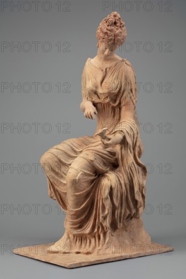 Figurine of a woman sitting on a rock with a flower in her hand. Woman sitting on a rock with a flower in her hand, towards which she bows her head. She is dressed in a thin chiton fastened at the shoulders and tied high under the breasts; a pleated cloak is thrown over her knees and left forearm. Hair is combed upwards. Flat quadrangular base. 4th century BC, Myrina. Figurine; height 31.5 cm, width 12.5 cm, depth 18 cm, weight 0.78 kg., A seated terracotta figure of a woman with a textured hairstyle and downcast face, wearing draped garments with flowing folds and gathered sleeves, one arm extended with an open hand and the other bent at the elbow, the sculpture showing a warm reddish-brown surface with lighter beige highlights, set against a smooth gray background on a flat rectangular base.