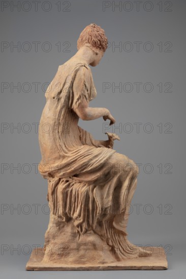 Figurine of a woman sitting on a rock with a flower in her hand. Woman sitting on a rock with a flower in her hand, towards which she bows her head. She is dressed in a thin chiton fastened at the shoulders and tied high under the breasts; a pleated cloak is thrown over her knees and left forearm. Hair is combed upwards. Flat quadrangular base. 4th century BC, Myrina. Figurine. Height 31.5 cm, width 12.5 cm, depth 18 cm, weight 0.78 kg., A seated terracotta figurine of a woman shown in profile on a rectangular base, rendered in a warm reddish-brown and pale beige clay color; she wears a long, draped garment with finely modeled folds and fringed sleeves, her short, textured hair is close to the head, and one arm is extended forward with a delicate hand above a small animal resting on her knee; the base and the seat beneath her show rough, sculpted rock-like texture in similar earthy tones, and the background is a smooth neutral gray.