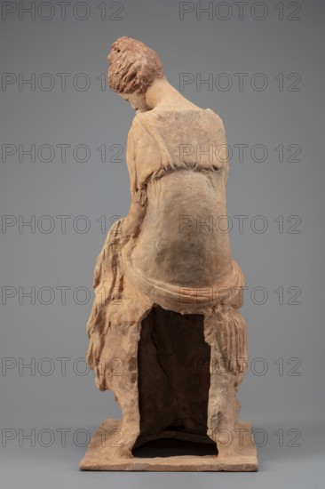Figurine of a woman sitting on a rock with a flower in her hand. Woman sitting on a rock with a flower in her hand, towards which she bows her head. She is dressed in a thin chiton fastened at the shoulders and tied high under the breasts; a pleated cloak is thrown over her knees and left forearm. Hair is combed upwards. Flat quadrangular base. 4th century BC, Myrina. Figurine; height 31.5 cm, width 12.5 cm, depth 18 cm, weight 0.78 kg., Flowers; female figurines., A sculpted figure viewed from behind, seated with a downward-tilted head and short textured hair, rendered in earthy tones of terracotta, brown, beige, and muted orange; the figure wears a draped garment with visible folds and gathered details, and rests on a hollowed base with rough, irregular edges that reveal a dark interior; the background is a smooth, neutral gray.