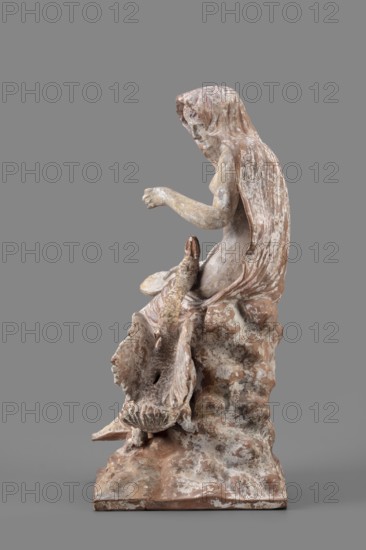 Figurine of Leda with a Swan, A woman sits on a rock and feeds a swan standing next to her. She is dressed in a cloak thrown over her head and back, falling along her body and completely enveloping her legs while leaving the rest of her body exposed. In her right hand, resting on her knees, she holds a platter; with her left hand, bent at the elbow and extended, she holds it above the swan, which rests on her exposed hip with its head extended toward her. The woman bows her head and looks at the swan. The swan has outstretched wings. The woman's sandaled feet do not rest on the rock but dangle. The square base forms one with the rock. Cloak painted pink. Inscription/note: Maria Bernhard, 01.06.1942. Date: mid-3rd century BC. Findspot/type: Myrina. Object type: figurine. Dimensions: height 24 cm, width 15 cm, depth 12 cm. Weight: 0.56 kg. Subjects/themes: Leda mythology, swans, figurines of women., A weathered sculpture of a seated figure draped in a hooded cloak, shown in profile again...