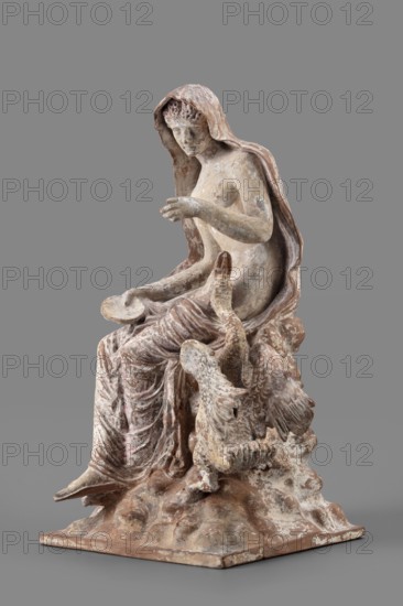 Figurine of Leda with a Swan, A woman sits on a rock and feeds a swan standing next to her. She is dressed in a cloak thrown over her head and back that falls along her body, completely enveloping her legs while leaving the rest of her body exposed. In her right hand, resting on her knees, she holds a platter; with her left arm bent and extended she holds it above the swan, which rests on her exposed hip with its head extended toward her. The woman bows her head and looks at the swan. The swan has outstretched wings. The woman's sandaled feet do not rest on the rock but dangle. The square base forms one with the rock. Cloak painted pink. Mid-3rd century BC, Myrina; terracotta figurine; height 24 cm, width 15 cm, depth 12 cm, weight 0.56 kg., A weathered sculptural figure of a seated person draped in a hooded cloak and flowing garments, rendered in muted tones of beige, tan, brown, and off-white with areas of pale pinkish and grayish patina; the figure's exposed torso and arm show subtl...
