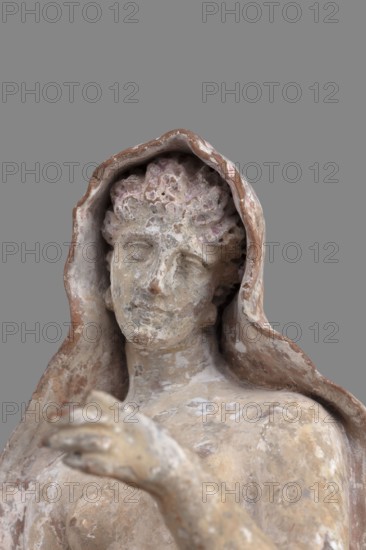 Figurine of Leda with a Swan, A woman sits on a rock and feeds a swan standing next to her. She is dressed in a cloak that, thrown over her head and back, falls along her body, completely enveloping her legs while leaving the rest of her body exposed. In her right hand, resting on her knees, she holds a platter. With her left arm, bent at the elbow and extended, she holds it above the swan, which rests on her exposed hip, its head extended towards her. The woman bows her head and looks at the swan. The swan has outstretched wings. The woman's sandaled feet do not rest on the rock but dangle. The square base forms one with the rock. Cloak painted pink. Mid-3rd century BC, Myrina. Terracotta figurine; height 24 cm, width 15 cm, depth 12 cm, weight 0.56 kg., A close-up of a weathered statue of a robed figure against a gray background showing a worn face with closed eyes and a subtle expression, curly textured hair partially covered by a hooded cloak, an outstretched arm and hand in the fo...