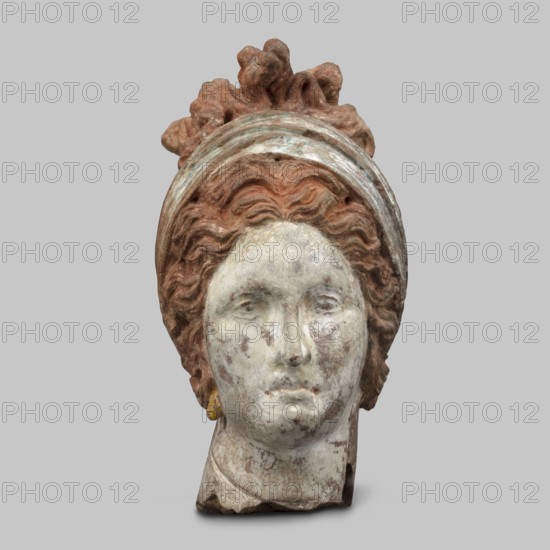 Head of a woman. Female head, fragment of a larger figurine. The head is tilted slightly to the right. The face is narrow, long. The straight nose forms a single plane with the forehead. Large, almond-shaped eyes are deeply set; near the nose, both eyelids are modeled. Eyebrow arches slope downward at the outside. Forehead triangular. Hair completely covers the ears, revealing only the earrings. Hair parted in the middle, forms waves at the back and is combed upwards, creating curls on the crown. Hair held in a wide ribbon that runs from the back to the front, crosses back and is tied. Polychrome perfectly preserved: face white with strong pink, traces of red on the lips; hair brown; scarf white and blue. 4th century BC, Tanagra Greece. Terracotta figurine. Height 13 cm, width 7 cm, depth 9.5 cm, weight 0.31 kg., A carved head sculpture shown against a plain light gray background, with a weathered face painted in pale off-white and gray tones and patches of darker discoloration, framed...