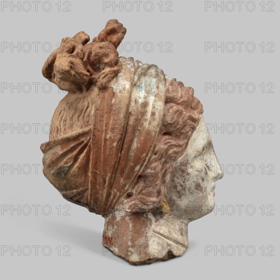 Head of a woman. Female head, fragment of a larger figurine. The head is tilted slightly to the right. The face is narrow, long. The straight nose forms a single plane with the forehead. The large, almond-shaped eyes are deeply set; near the nose, they have modeled both eyelids. The arches of the eyebrows slope downward on the outside. The forehead is triangular. The hair completely covers the ears, revealing only the earrings. The hair, parted in the middle, forms waves at the back and is combed upwards, creating curls on the crown. The hair is held in a wide ribbon, which, when worn from the back, runs to the front, where it crosses back and is tied. The polychrome is perfectly preserved: face white with strong pink tones and traces of red on the lips; hair brown; scarf white and blue. 4th century BC, Tanagra Greece. Figurine. Height 13 cm, width 7 cm, depth 9.5 cm, weight 0.31 kg., A weathered sculpted head in profile facing right, showing worn carved hair and a draped head covering...