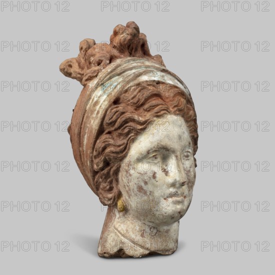 Head of a woman. Female head, fragment of a larger figurine. The head is tilted slightly to the right. The face is narrow, long. The straight nose forms a single plane with the forehead. The large, almond-shaped eyes are deeply set; near the nose they have modeled both eyelids. The arches of the eyebrows slope downward on the outside. The forehead is triangular. The hair completely covers the ears, revealing only the earrings. The hair, parted in the middle, forms waves at the back and is combed upwards, creating curls on the crown. The hair is held in a wide ribbon, which, when worn from the back, runs to the front, where it crosses back and is tied. The polychrome is perfectly preserved: the face is white, very pink, with traces of red on the lips; the hair is brown; the scarf is white and blue. 4th century BC, Tanagra Greece. Terracotta figurine; height 13 cm, width 7 cm, depth 9.5 cm, weight 0.31 kg., A sculpted head of a person rendered in weathered stone tones and reddish-brown m...