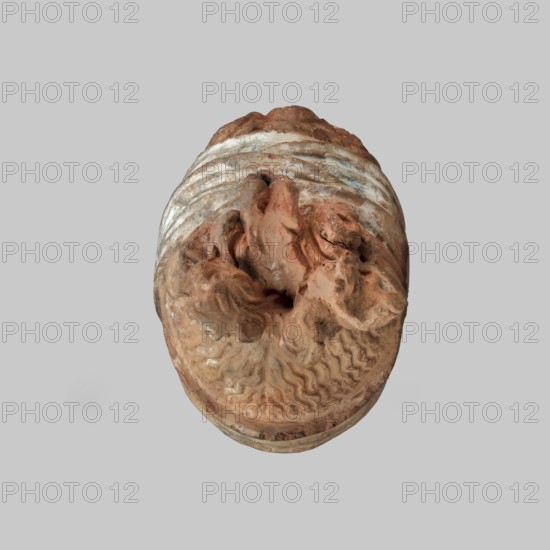 Head of a woman, Female head, fragment of a larger figurine. The head is tilted slightly to the right. The face is narrow, long. The straight nose forms a single plane with the forehead. Large almond-shaped eyes are deeply set; near the nose both eyelids are modeled. The arches of the eyebrows slope downward on the outside. The forehead is triangular. The hair completely covers the ears, revealing only the earrings. The hair, parted in the middle, forms waves at the back and is combed upwards, creating curls on the crown. The hair is held in a wide ribbon that, when worn from the back, runs to the front, crosses back and is tied. The polychrome is well preserved: the face is white with pink tones and traces of red on the lips; the hair is brown and the scarf white and blue. 4th century BC, Tanagra Greece. Terracotta figurine; height 13 cm, width 7 cm, depth 9.5 cm, weight 0.31 kg., An oval-shaped sculpted object viewed from above showing textured, irregular surfaces with carved, wavy p...