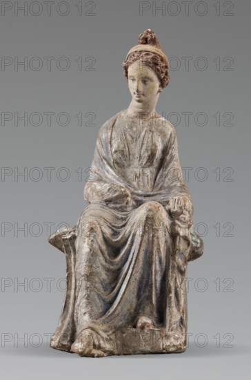 Figurine of a woman. A girl sitting in a chair is dressed in a soft white chiton over which a blue, in some places lilac, cloak is thrown, which opens at the front. She rests her left hand on the chair cushion, while her right rests on her knees and is covered by the cloak. Her brown hair is parted at the front, combed upwards and girded. The full chair with forward-curved legs has a narrow and low backrest; the seat cushion is blue on top and red underneath. No base. Early 3rd century BC, Tanagra Greece. Terracotta figurine, height 16 cm, width 8 cm, depth 10 cm, weight 0.25 kg., A seated statue of a robed female figure with detailed drapery, showing a textured surface and worn paint in shades of beige, gray, brown, muted blue, and hints of reddish-brown on the hair and headband; the figure has curled hair pulled into a topknot, a calm facial expression, and hands resting on the lap and armrest; the robe forms flowing folds over the legs and down to the base, and the figure sits on a ...