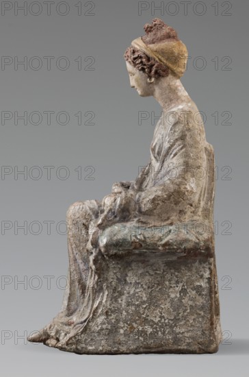 Figurine of a woman. A girl sitting in a chair is dressed in a soft white chiton over which a blue, in some places lilac, cloak is thrown, which opens at the front. She rests her left hand on the chair cushion, while her right rests on her knees and is covered by the cloak. Her brown hair is parted at the front, combed upwards and girded. The full chair with forward-curved legs has a narrow and low backrest; the seat cushion is blue on top and red underneath. No base. Early 3rd century BC, Tanagra Greece. Terracotta figurine; height 16 cm, width 8 cm, depth 10 cm, weight 0.25 kg., A seated figure sculpted in a weathered material shown in profile facing left, wearing a head covering with textured hair visible at the front and a rounded bun at the back, the head covering and hair showing shades of brown and yellow; the face and exposed neck are light beige with some darker discoloration; the garment draped over the body has folds and a worn surface in tones of gray and off-white with hin...