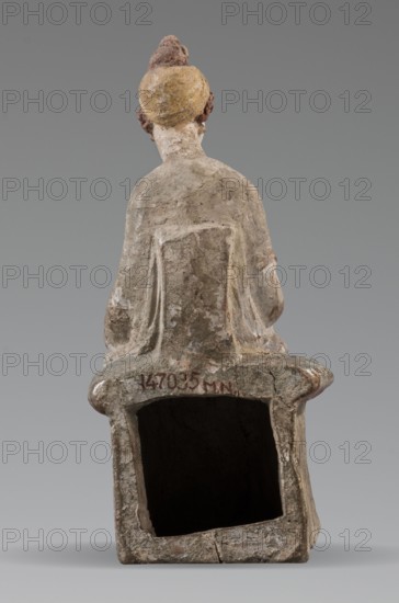 Figurine of a woman. A girl sitting in a chair is dressed in a soft white chiton over which a blue, in some places lilac, cloak is thrown, which opens at the front. She rests her left hand on the chair cushion, while her right rests on her knees and is covered by the cloak. Her brown hair is parted at the front, combed upwards and girded. The full chair with forward-curved legs has a narrow and low backrest; the seat cushion is blue on top and red underneath. No base. Early 3rd century BC, Tanagra Greece. Terracotta figurine; height 16 cm, width 8 cm, depth 10 cm, weight 0.25 kg., A rear view of a seated human figure sculpture with a weathered surface showing a palette of gray, beige, brown, ochre, and muted yellow; the figure has a rounded headpiece with brown and yellow tones and short hair colored brown, a neck and back with grayish-beige textures, and draped clothing suggested by carved folds in gray and beige; the seat and base share the same aged, mottled gray and brown appearanc...