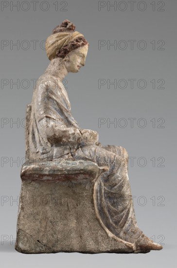 Figurine of a woman. A girl sitting in a chair is dressed in a soft white chiton over which a blue, in some places lilac, cloak is thrown, which opens at the front. She rests her left hand on the chair cushion, while her right rests on her knees and is covered by the cloak. Her brown hair is parted at the front, combed upwards and girded. The full chair with forward-curved legs has a narrow and low backrest; the seat cushion is blue on top and red underneath. No base. Early 3rd century BC, Tanagra Greece. Terracotta figurine, height 16 cm, width 8 cm, depth 10 cm, weight 0.25 kg., A seated sculpture of a robed figure shown in profile, with a headwrap and hair detail, sitting on a block-like seat; the figure's robe has visible folds and drapery, and the hands rest in the lap. The surface shows weathering and wear with areas of rough texture and small cracks. Visible colors include muted beige and off-white on the skin and seat, pale yellow and brown tones in the headwrap and hair, faded...