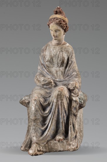 Figurine of a woman. A girl sitting in a chair is dressed in a soft white chiton over which a blue, in places lilac, cloak is thrown, opening at the front. She rests her left hand on the chair cushion, while her right rests on her knees and is covered by the cloak. Her brown hair is parted at the front, combed upwards and girded. The full chair with forward-curved legs has a narrow, low backrest; the seat cushion is blue on top and red underneath. No base. Early 3rd century BC, Tanagra Greece. Terracotta figurine, height 16 cm, width 8 cm, depth 10 cm, weight 0.25 kg., A seated sculpted figure wearing a long draped garment with visible folds and a shawl, with curly hair gathered under a headband and a small raised element atop the head; the figure’s hands rest in the lap and on the side of the seat, and the feet extend forward from beneath the drapery. The sculpture shows a weathered surface with areas of muted beige, off-white, gray, brown, reddish tones, and traces of faded blue. The...
