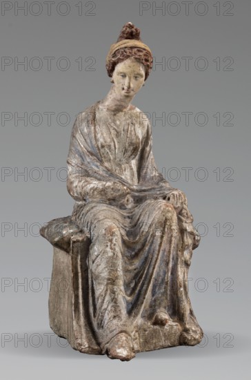 Figurine of a woman. A girl sitting in a chair is dressed in a soft white chiton over which a blue, in places lilac, cloak is thrown, opening at the front. She rests her left hand on the chair cushion, while her right rests on her knees and is covered by the cloak. Her brown hair is parted at the front, combed upwards and girded. The full chair with forward-curved legs has a narrow, low backrest; the seat cushion is blue on top and red underneath. No base. Early 3rd century BC, Tanagra Greece. Terracotta figurine; height 16 cm, width 8 cm, depth 10 cm, weight 0.25 kg., A seated terracotta figure wearing a draped garment with visible folds, shown from front with a slightly tilted head and a neutral facial expression; the hair is styled up and topped with a wrapped band and a small knot, and the hands rest lightly on the lap and beside the knee; the seat is a simple block with a padded cushion area; the surface shows worn paint and weathered texture with colors including muted beige, gra...