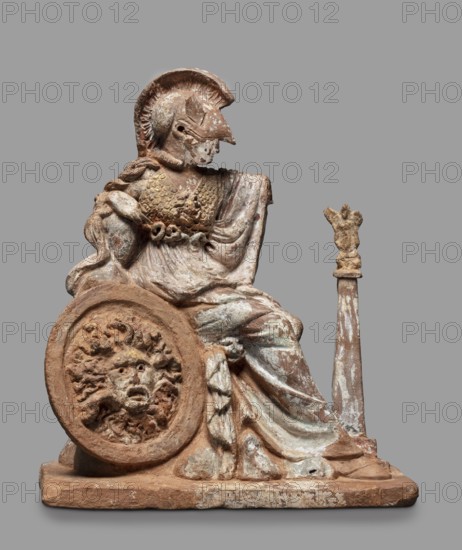 Athena figurine, Classical, 4th century BC, Greece; subject: Athena and Nike Greek deities; type: figurine; materials not specified; dimensions: height 16.5 cm, width 13.5 cm, depth 7.8 cm; weight 0.35 kg; inscription/marking: Froehner, V., Ant., A weathered terracotta figure seated on a rectangular base wearing a helmet with a crest and a draped cloak, showing textured armor on the torso and a braided detail at the shoulder; the figure holds a round shield resting against the base with a raised grotesque face motif, and beside the figure stands a tall staff topped with a small sculpted ornament. The sculpture exhibits earthy brown and reddish tones with patches of white and pale green paint or patina, and areas of darker brown where the material is exposed.