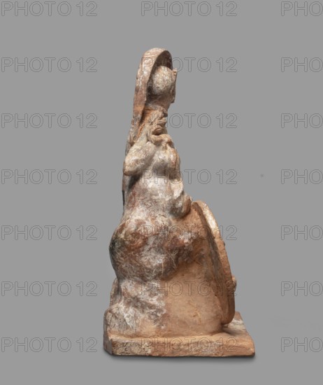 Athena figurine, Classical Greek, 4th century BC, Greece; depicts Athena with Nike, terracotta, figurine; height 16.5 cm, width 13.5 cm, depth 7.8 cm, weight 0.35 kg., A weathered sculpture of a seated figure shown in profile against a plain gray background, with a rough textured surface in shades of beige, tan, brown, and off-white; the figure wears a rounded head covering or halo and has long flowing hair or drapery down the back, an arm raised toward the chest, and another arm resting near a rounded shield- or disk-like object at the side; the base is rectangular and shows the same worn coloration and textured stone appearance.