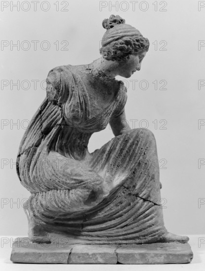 Terracotta figurine of a girl throwing dice. Figurine of a crouching girl throwing dice: the weight rests on the left leg, bent at a right angle. She wears a long, finely pleated, sleeveless chiton. Hair tied in a bun with a wide headband kredemnos. Earring visible in left ear. Entire figurine rests on a rectangular base. Preservation: missing right arm; glued in several places head, left arm from 2 fragments, robe, base from 3 fragments. Part of base filled with clay and plaster. 4th–3rd century BC, Tanagra Greece. Terracotta; height 13 cm, width 9 cm, depth 4.5 cm., A grayscale photograph shows a sculpted figure seated on a low rectangular base, depicted in profile with head turned slightly downward; the figure wears a draped garment with carved folds and textured lines, and hair styled in a bun with visible curls; the sculpture surface appears weathered with visible cracks and rough patches; the base and lower part of the garment show layered horizontal grooves and fracture lines; t...
