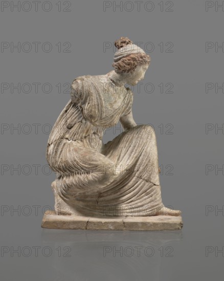 Terracotta figurine of a girl throwing dice. Figurine of a crouching girl throwing dice: weight of the body rests on the left leg, bent at a right angle. She wears a long, finely pleated, sleeveless chiton. Hair tied in a bun with a wide headband kredemnos. Earring visible in left ear. Entire figurine rests on a rectangular base. Preservation: missing right arm; glued in several places head; left arm from 2 fragments; robe; base from 3 fragments. Part of the base filled with clay and plaster. Date: 4th–3rd century BC. Place of origin: Tanagra Greece. Material: terracotta. Type: mold-made Tanagrian-style figurine. Dimensions: height 13 cm, width 9 cm, depth 4.5 cm. Creator: unknown., A sculpted figure of a seated woman in a flowing draped garment, shown in profile facing right, with hair gathered in a bun; the surface shows worn textures and layered folds in the clothing, and the piece rests on a rectangular base. The visible colors include shades of gray, beige, off-white, light brown,...