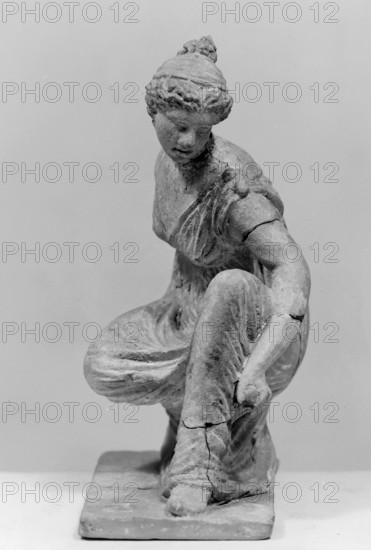 Terracotta figurine of a girl throwing dice. Figurine of a crouching girl throwing dice: the weight of the body rests on the left leg, bent at a right angle. She wears a long, finely pleated, sleeveless chiton. Hair tied in a bun with a wide headband kredemnos. Earring visible in left ear. Entire figurine rests on a rectangular base. Preservation: missing the right arm; glued in several places head, left arm from 2 fragments, robe, base from 3 fragments; part of the base filled with clay and plaster. Date: 4th–3rd century BC. Origin: Tanagra Greece. Material: terracotta. Type: figurine. Dimensions: height 13 cm, width 9 cm, depth 4.5 cm. Maker: unknown., A grayscale photograph of a sculpted figure of a seated woman on a rectangular base, shown from a slightly elevated frontal angle; the figure wears a draped garment with visible folds and appears to be adjusting or holding the drapery near a bent knee, with one arm extended downward and the other arm resting near the torso; the head is...